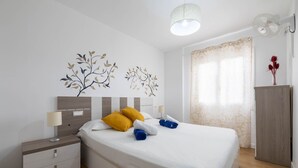 A neatly made bed with white bedding, two pillows, and two rolled towels. There is a bedside table with a lamp, a wall-mounted artwork of branches, and a window with curtains. - Sol y Mar by Rentallorca (Can Picafort)