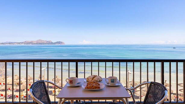 A balcony with a table set for two, overlooking a beach with umbrellas and lounge chairs. - Sol y Mar by Rentallorca (Can Picafort)