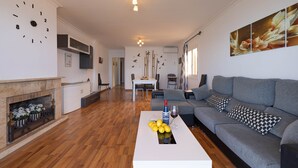 A modern living room with a fireplace, a sofa, a coffee table with lemons and a bottle, and a dining area in the background. - Sol y Mar by Rentallorca (Can Picafort)