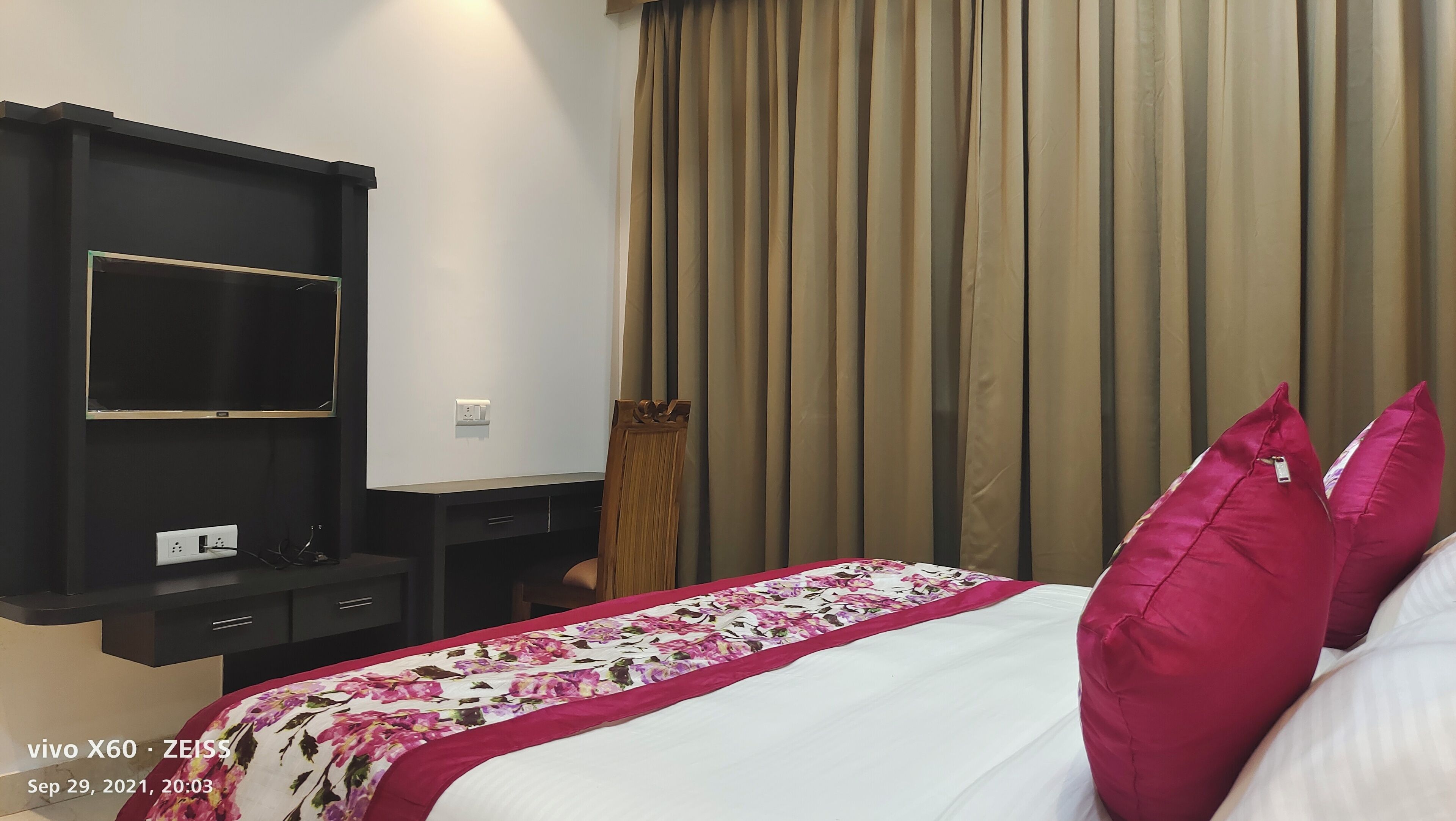 A hotel room with a bed, a TV on a stand, a desk with a computer, and a window with curtains.