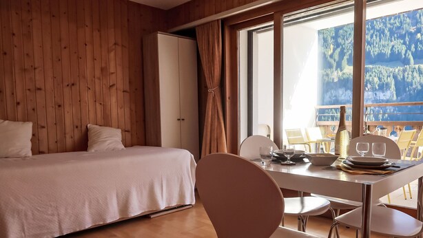 Une chambre avec un lit, une table à manger et des chaises, et une vue sur une forêt à travers de grandes fenêtres.