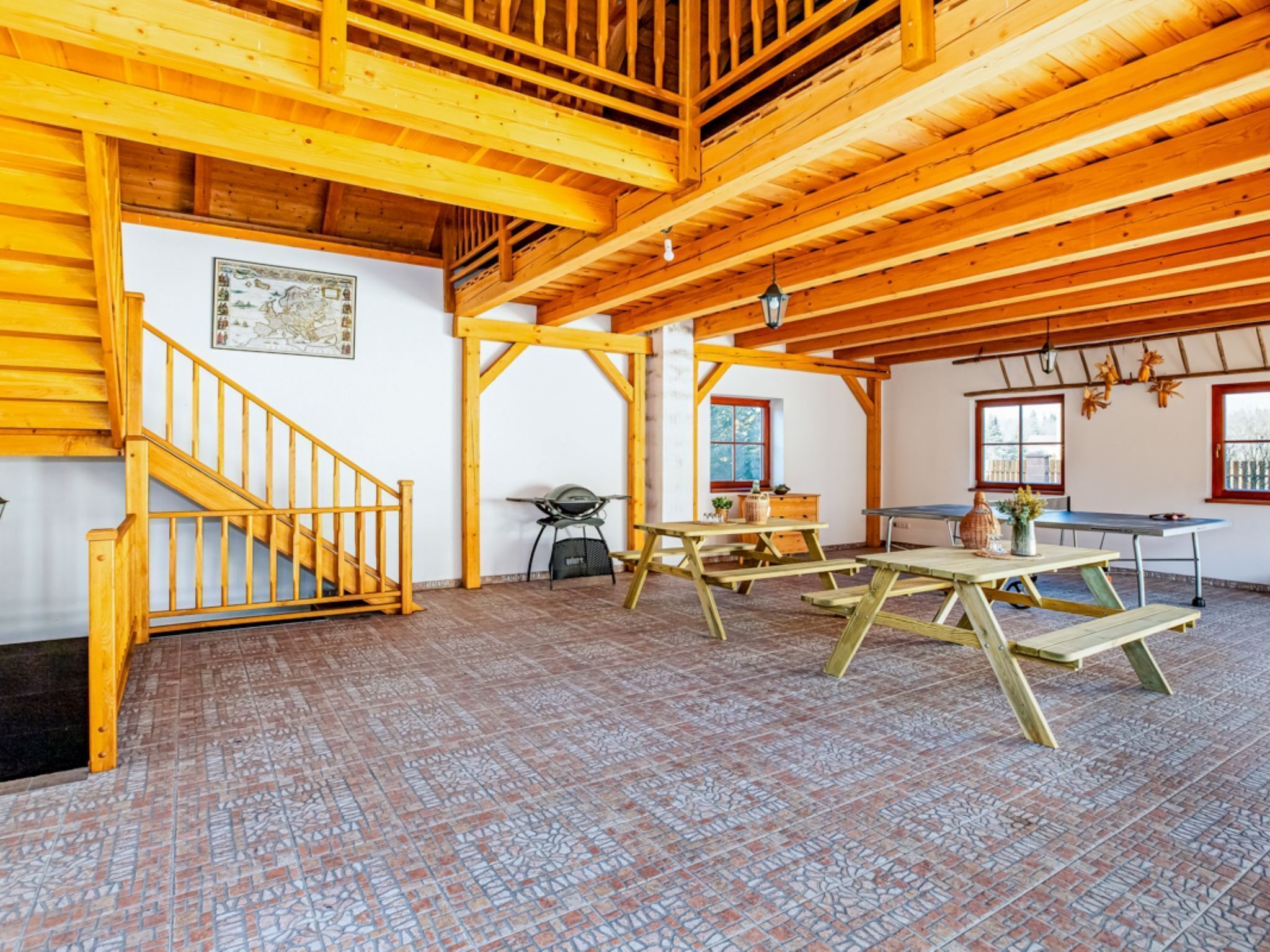 A spacious room with a high wooden ceiling, a wooden staircase, a table with chairs, and a window with a view of the outdoors.