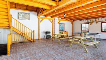 A spacious room with a high wooden ceiling, a wooden staircase, a table with chairs, and a window with a view of the outdoors.