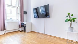A room with a large flat-screen TV mounted on the wall, a radiator, a desk, a chair, a potted plant, and a window with curtains. - Affordable Cozy 2BR Apt Near Stadthalle (Vienna)