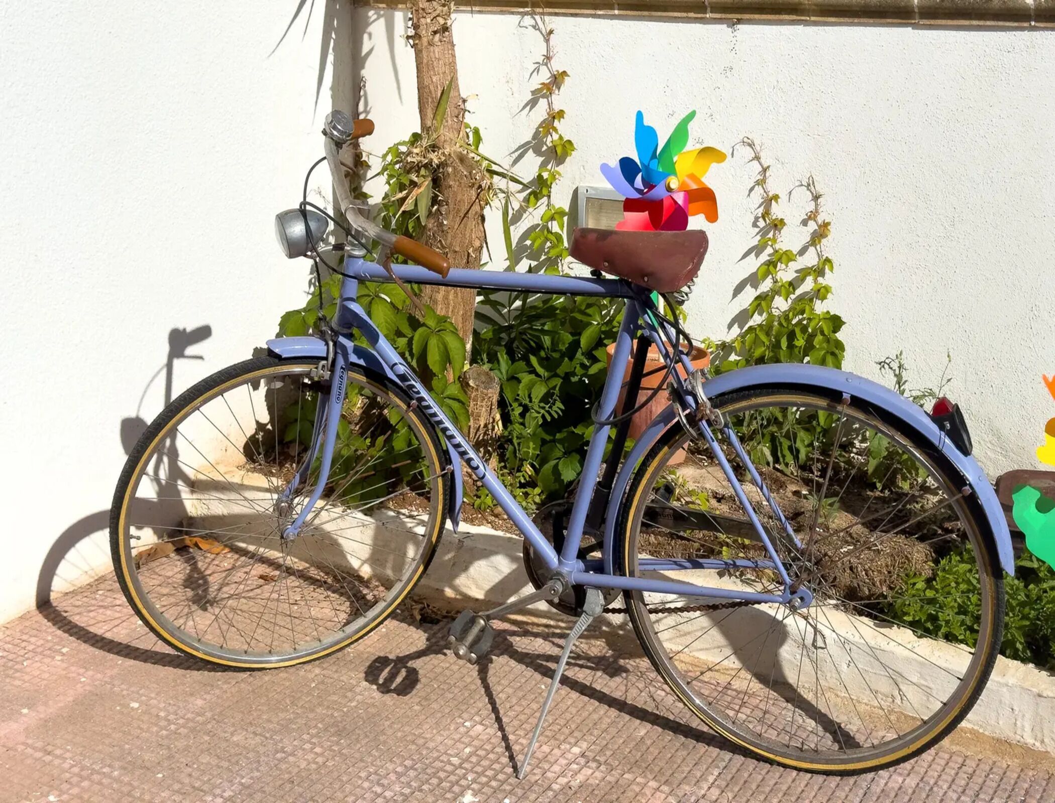 Een blauw fietsje met een mandje en bloemendecoratie, geparkeerd tegen een muur met groen.