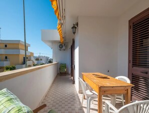 A balcony with a wooden table and white chairs, a green chair, a small table with a bowl, and a wall-mounted air conditioner. - Villetta Marinaci Porto Cesareo (Porto Cesareo)
