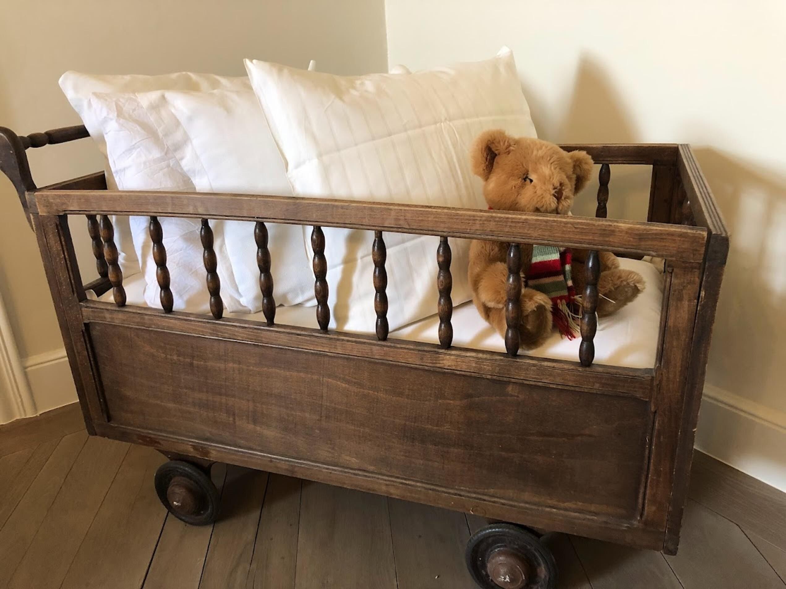 Una cuna de madera con un oso dentro, ruedas y almohadas blancas.