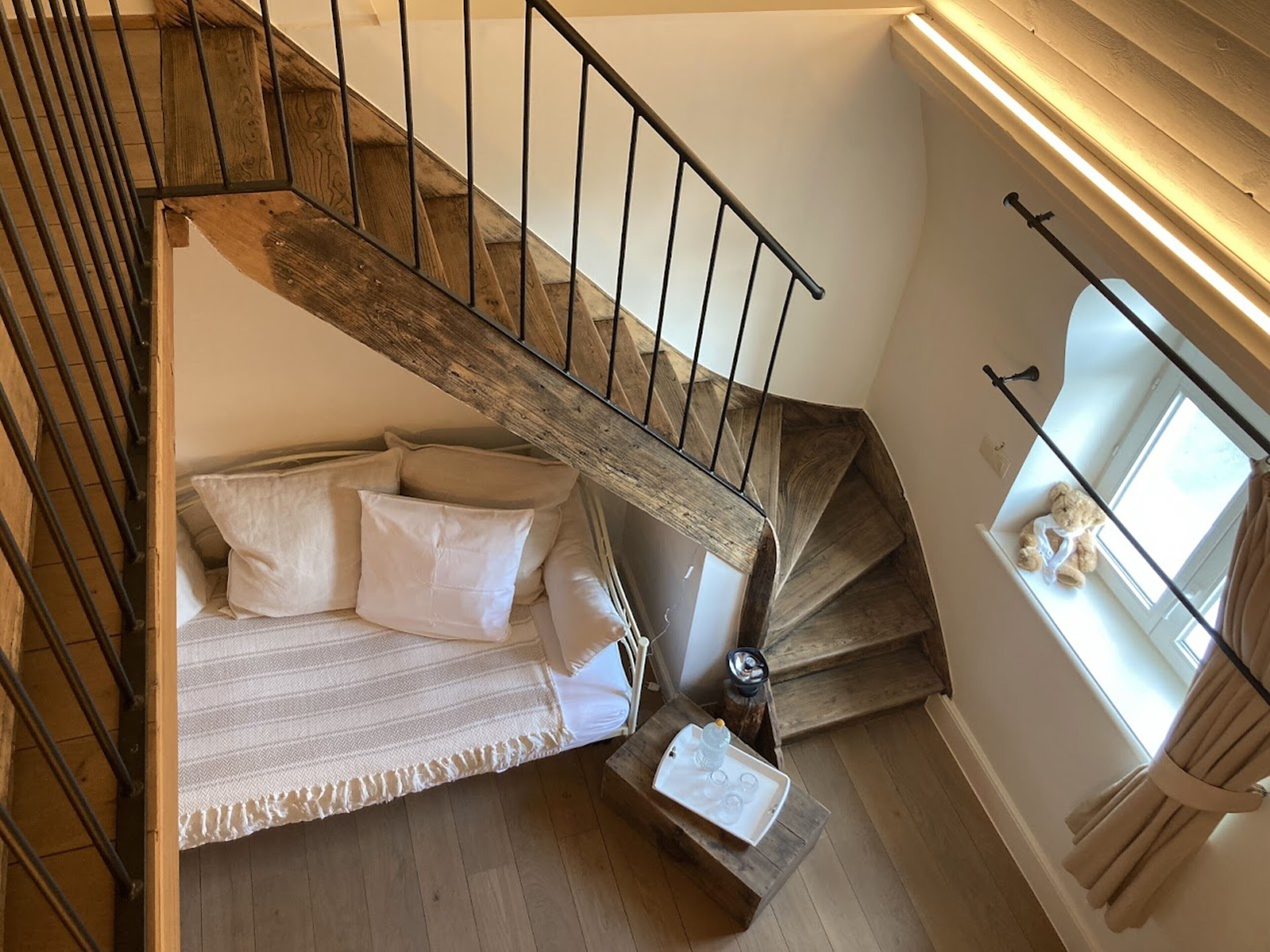 Un escalier avec des marches en bois et des rampes en métal mène à une chambre à coucher où se trouvent un lit, des oreillers et une table de chevet sur laquelle repose un récipient en verre.