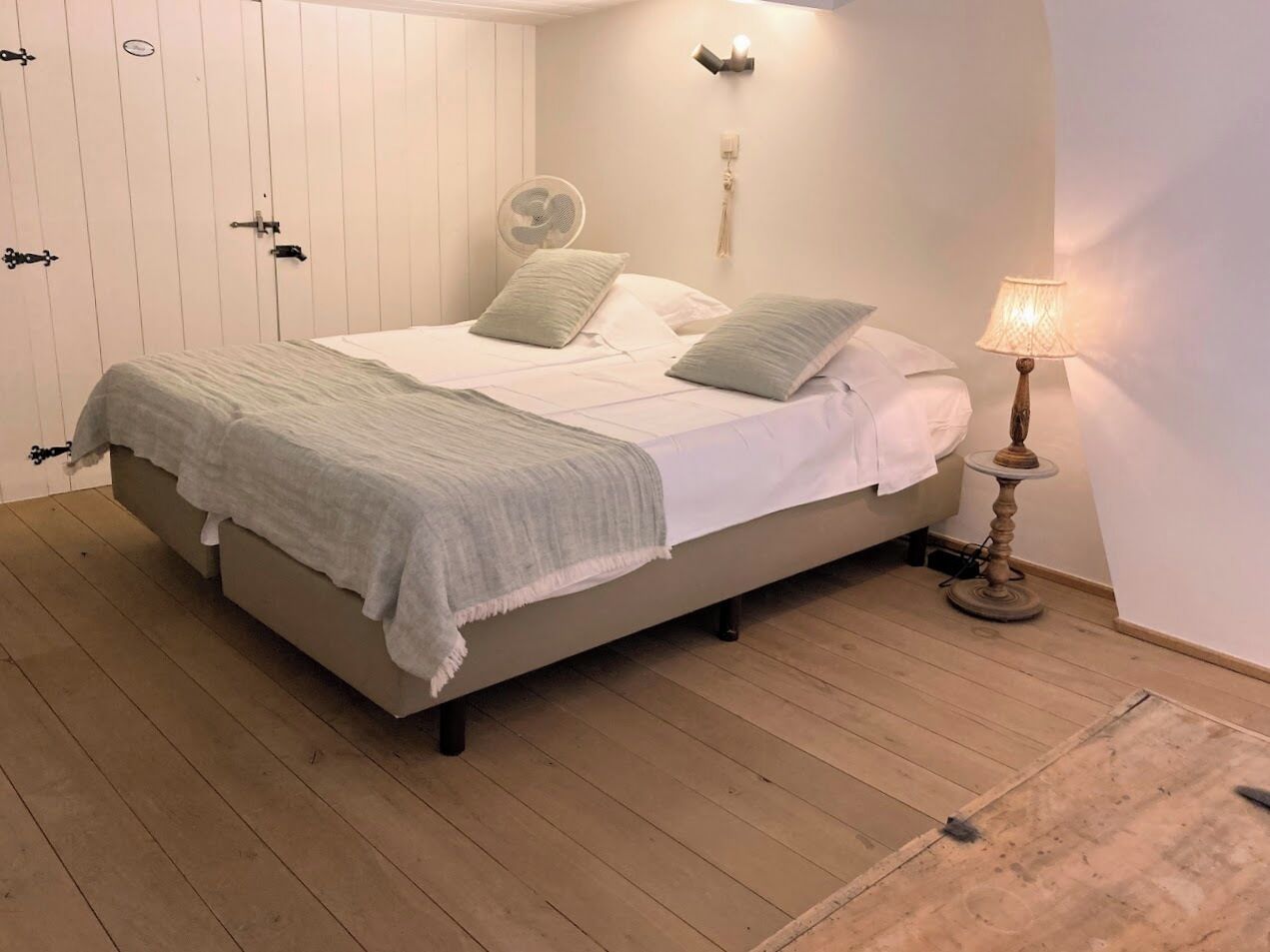 A bedroom with a bed, a bedside table with a lamp, a wall-mounted fan, and a wardrobe.