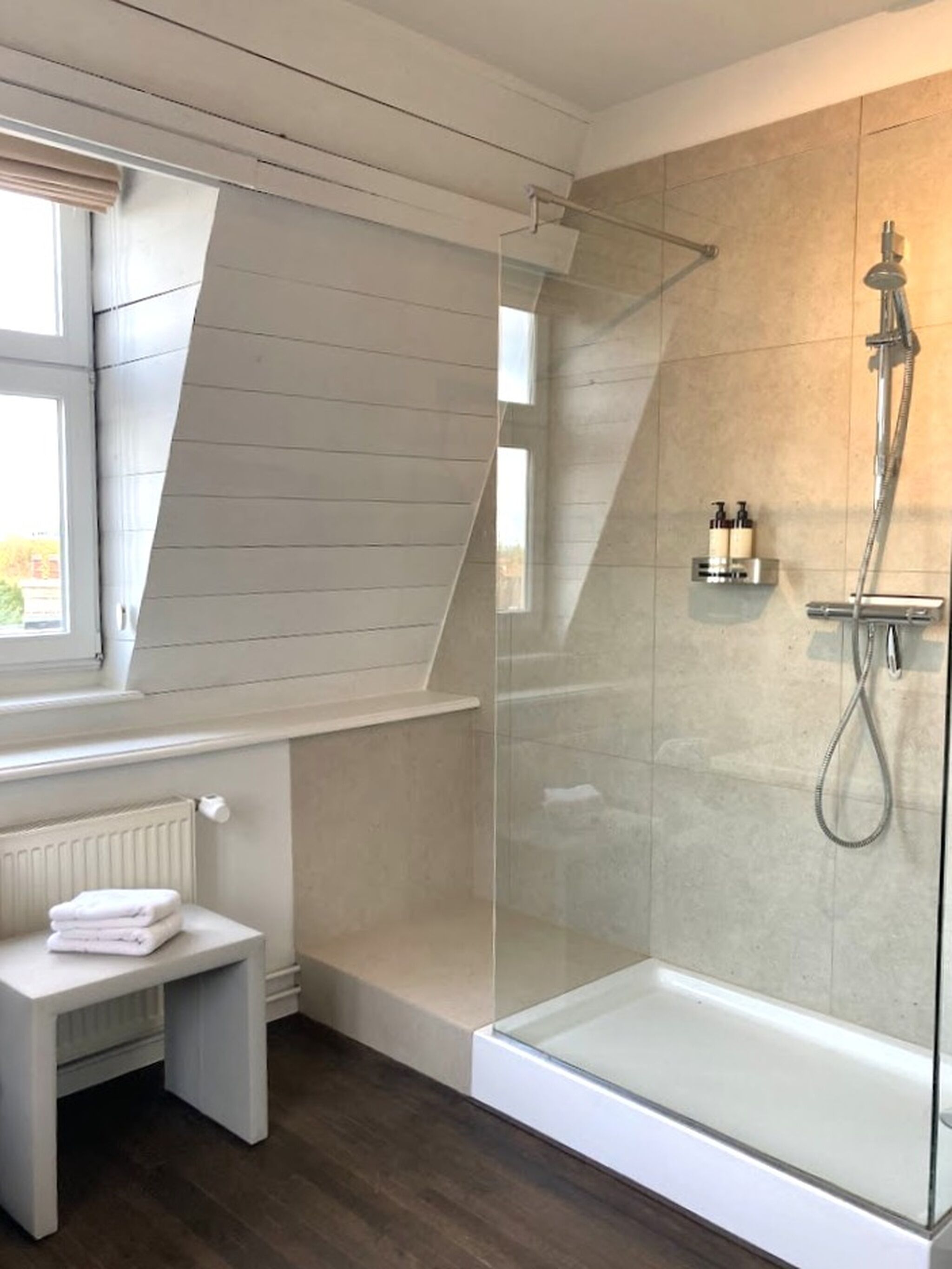 A modern bathroom with a glass-enclosed shower, a white bench with towels, and a window providing natural light.