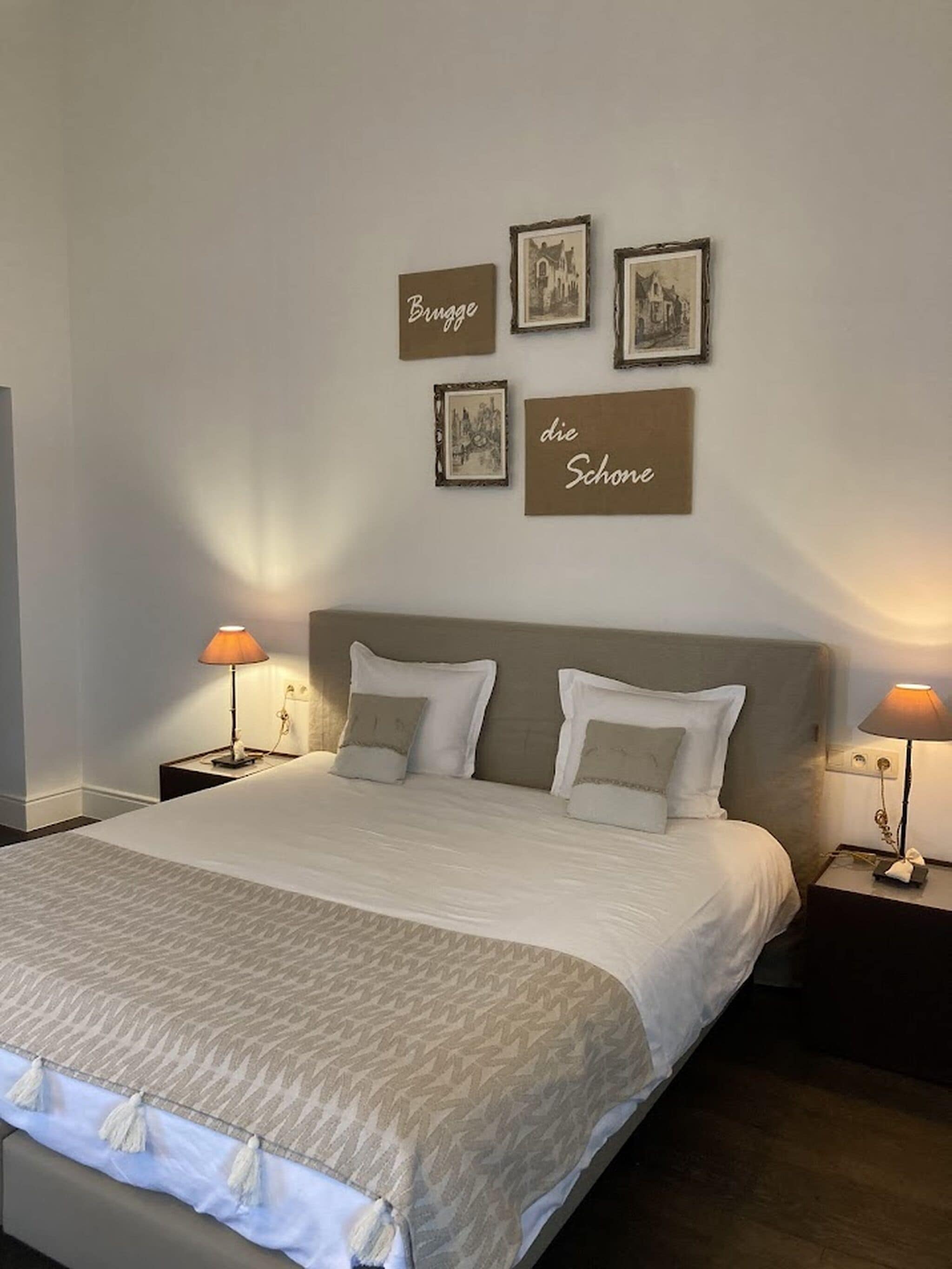Un lit bien fait, recouvert d’une couvre-lit à motifs et agrémenté de deux oreillers, est flanqué de deux tables de chevet sur lesquelles reposent des lampes. Au mur, on peut voir trois tableaux encadrés et deux panneaux avec du texte.