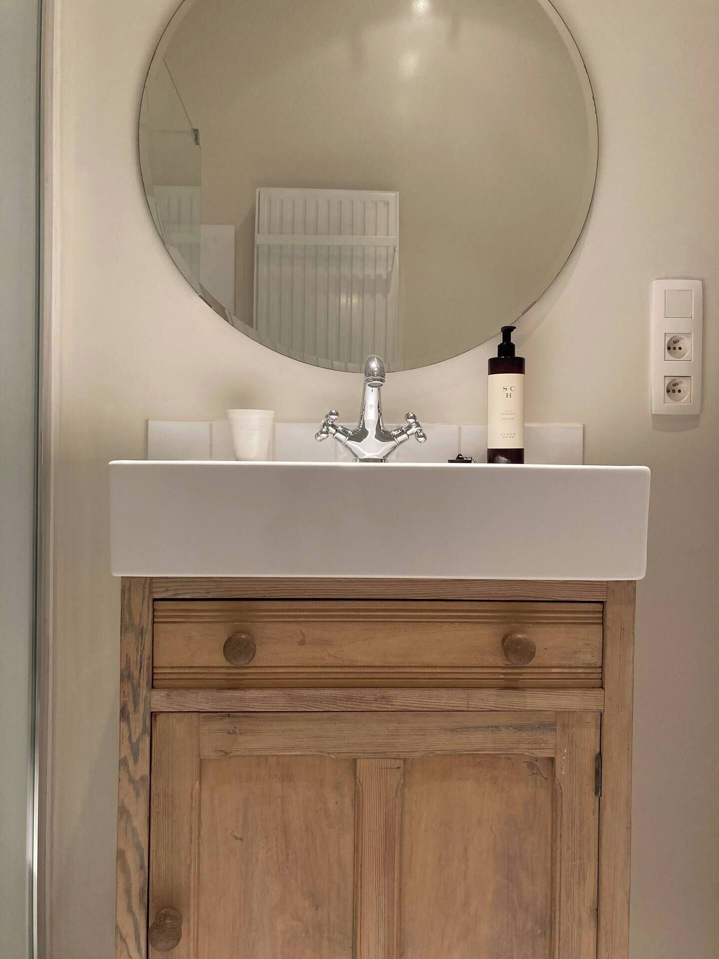 A bathroom with a round mirror, a white sink, and a wooden cabinet below.