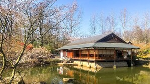 Il s’agit d’un bâtiment japonais traditionnel, avec une façade en bois, un toit recouvert de tuiles et une base en pierre, situé au bord d’un étang paisible dont l’eau reflète le paysage.