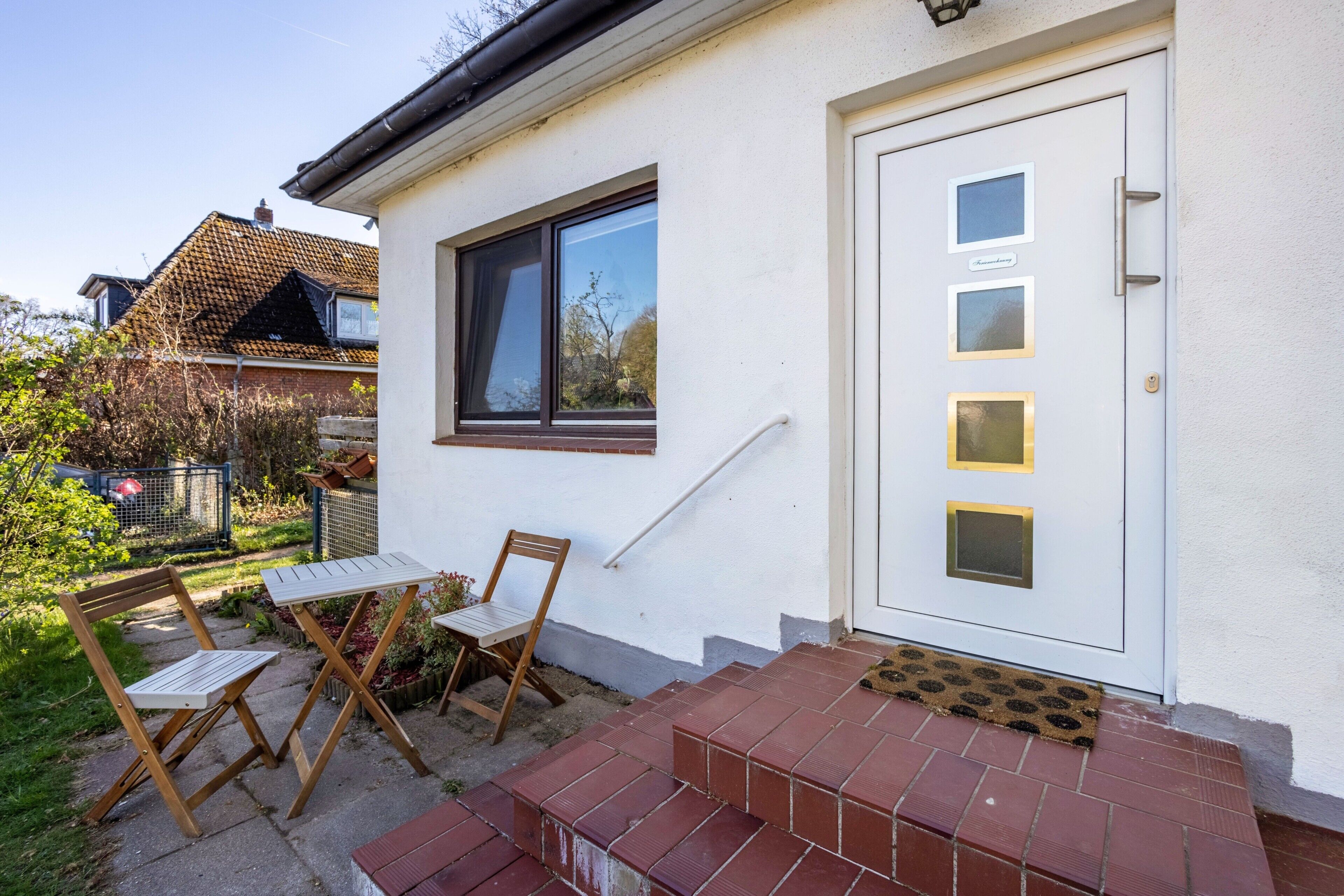 A white house with a white door, a window, and a small outdoor seating area with two wooden chairs and a table.