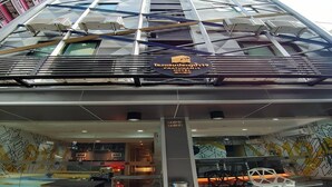 The exterior of the Pratunam Hotel with a modern glass facade, a visible restaurant area inside, and a sign indicating the hotel's name and location. - Affordable Single | Central Thailand Stay (Bangkok)