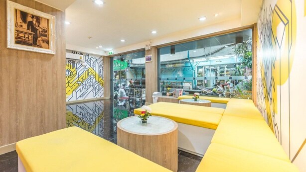 A modern interior with yellow seating, a glass wall, and a visible outdoor area with shops. - Affordable Single | Central Thailand Stay (Bangkok)