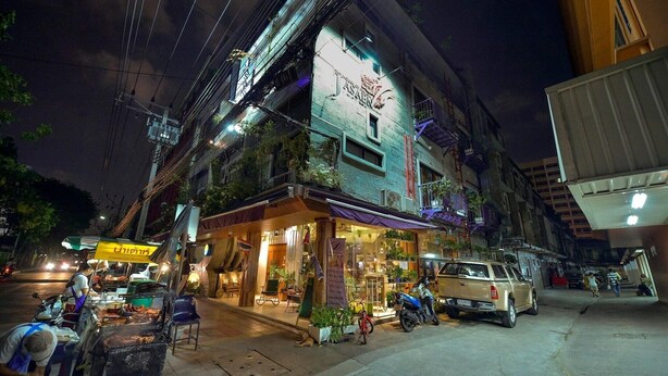 A street view at night with a building featuring a large illuminated sign, outdoor seating, and a parked motorcycle. - Solo Retreat in Style | Standard Single Stay (Bangkok)