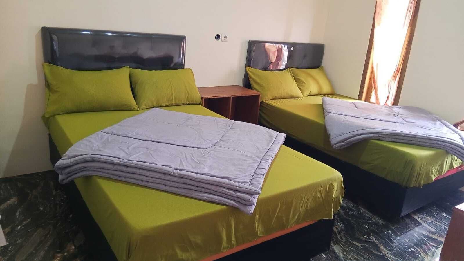 A hotel room with two beds, each with green bedding and a grey blanket. There is a wooden nightstand between the beds.