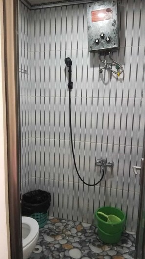 Une petite salle de bain carrelée, équipée d’une douche, de toilettes et d’un seau vert.