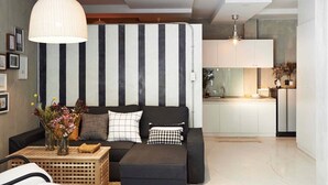 Ein modernes Wohnzimmer mit einer schwarz-weißen Streifenwand, einer Couch mit Kissen, einer Pendelleuchte und einer Küchenzeile im Hintergrund.