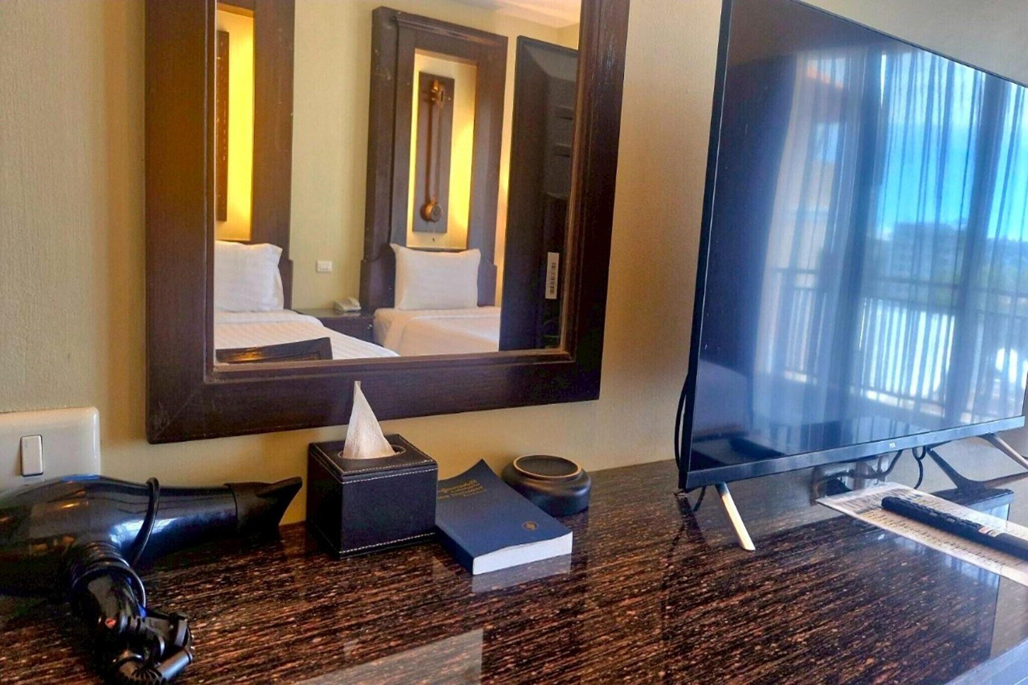 A hotel room with a large mirror, a flat-screen TV, a wooden bedside table, a book, a tissue box, and a remote control.
