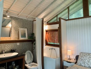 Un baño con lavamanos, inodoro y bañera. Hay un espejo sobre el lavamanos, un cuadro en la pared y una ventana con cortinas.
