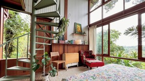 A room with a spiral staircase, wooden furniture, a colorful floral bedspread, and large windows offering a view of greenery. - Hilltop Retreat Beneath Mt. Batukaru (Selemadeg)