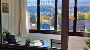 A room with a large window offering a view of a hilly landscape and a traditional house. There is a small table with a decorative plate and a chair. - Haus Sonnenschein an der Skipiste (Herrischried)