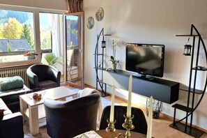 Ein modernes Wohnzimmer mit Fernseher, bequemen Sitzmöglichkeiten, einem kleinen Tisch und Blick ins Freie.