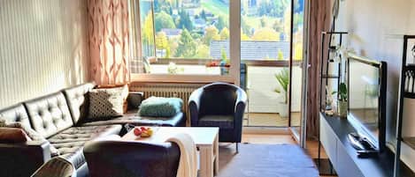 A living room with a sofa, armchairs, a coffee table, and a TV. There is a large window with a view of a green landscape.