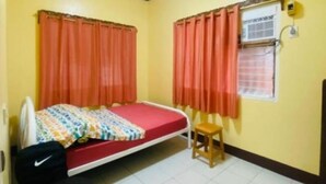 A small, single-bed room with a red mattress, a wooden stool, and two windows with orange curtains.
