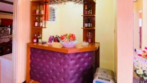 Zona de bar con mostrador acolchado color púrpura, estanterías con botellas y un pequeño arreglo floral.