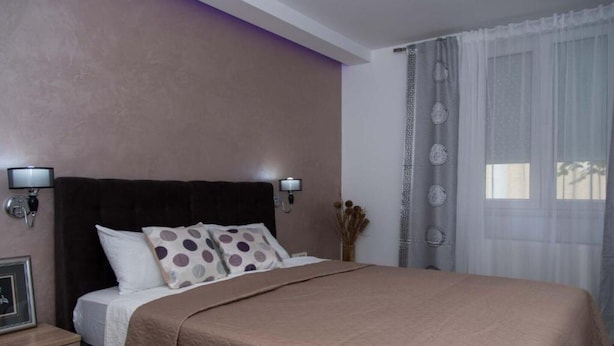 Un dormitorio ordenado con una cama grande, dos lámparas de noche, una mesita de noche y una ventana con cortinas traslúcidas.
