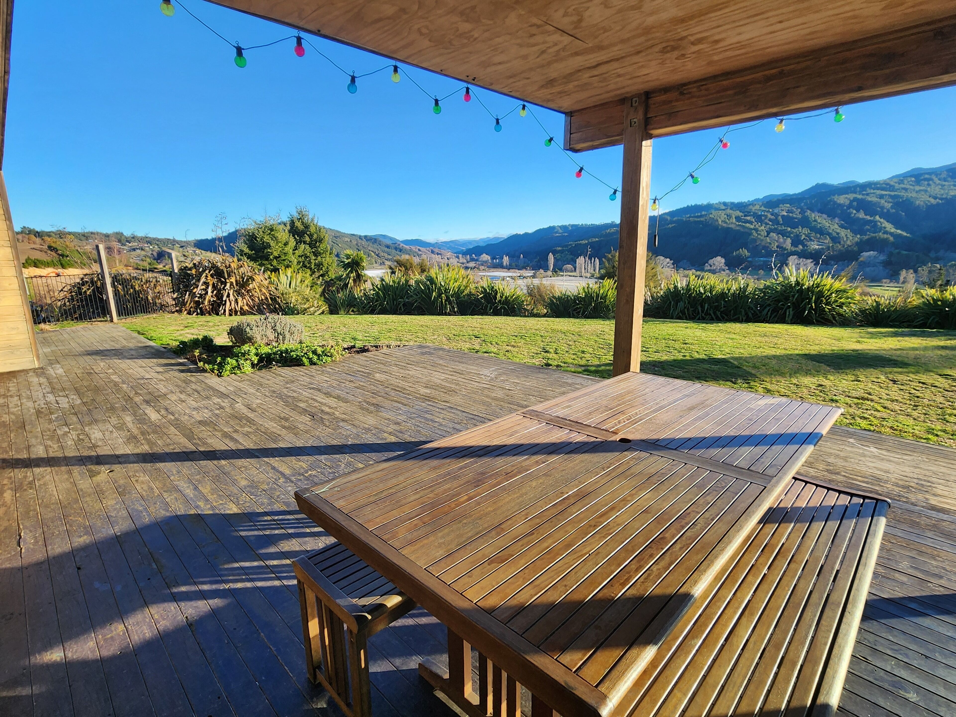 Eine Holzterrasse mit Tisch und Bänken, mit Blick auf eine Wiese und dahinter Berge.