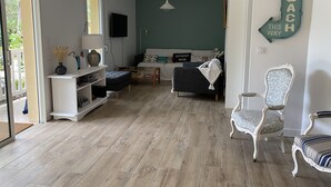Uma sala de estar moderna com sofá, apoio de pés e uma poltrona decorativa. Há uma televisão na parede e uma placa com a inscrição 'BEACH THIS WAY'.