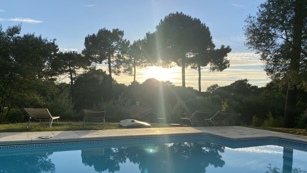 Piscina com espreguiçadeiras e um banco, rodeada por árvores e um céu limpo.