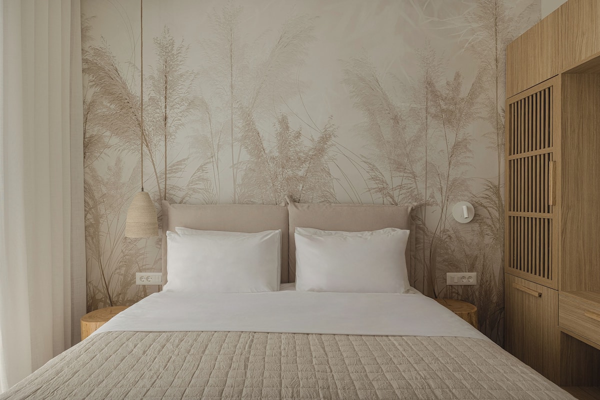 Un lit bien fait, recouvert d’une courtepointe, une table de chevet en bois avec une lampe, et un mur orné d’une fresque sur le thème de la nature.