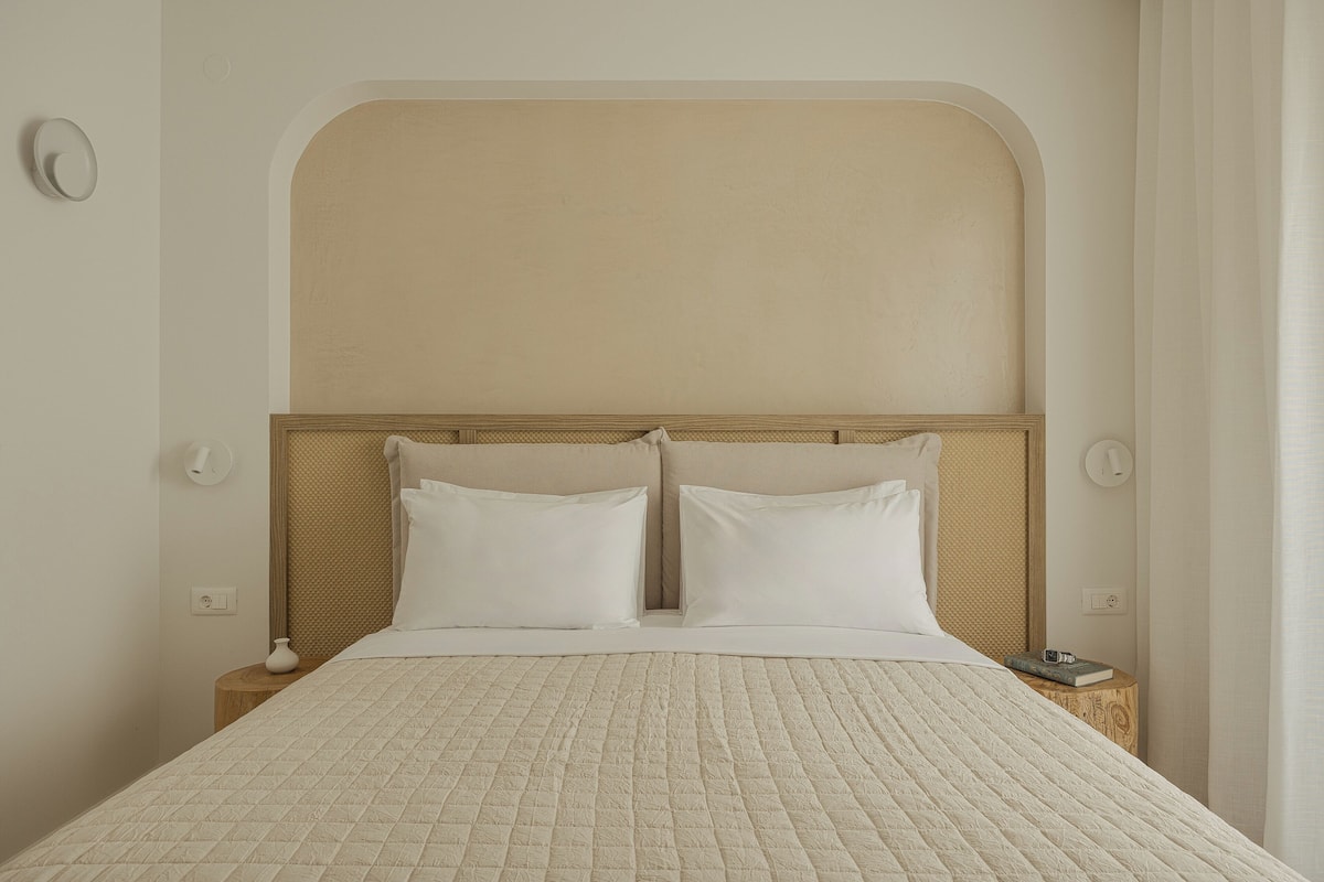 Un lit bien fait, avec une couvre-lit matelassé et deux oreillers, une tête de lit en bois et une table de chevet sur laquelle se trouvent un livre et un vase.