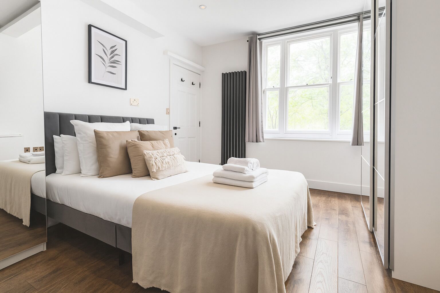 A modern bedroom with a large bed, a bedside table, a framed artwork, a radiator, and a window with curtains.