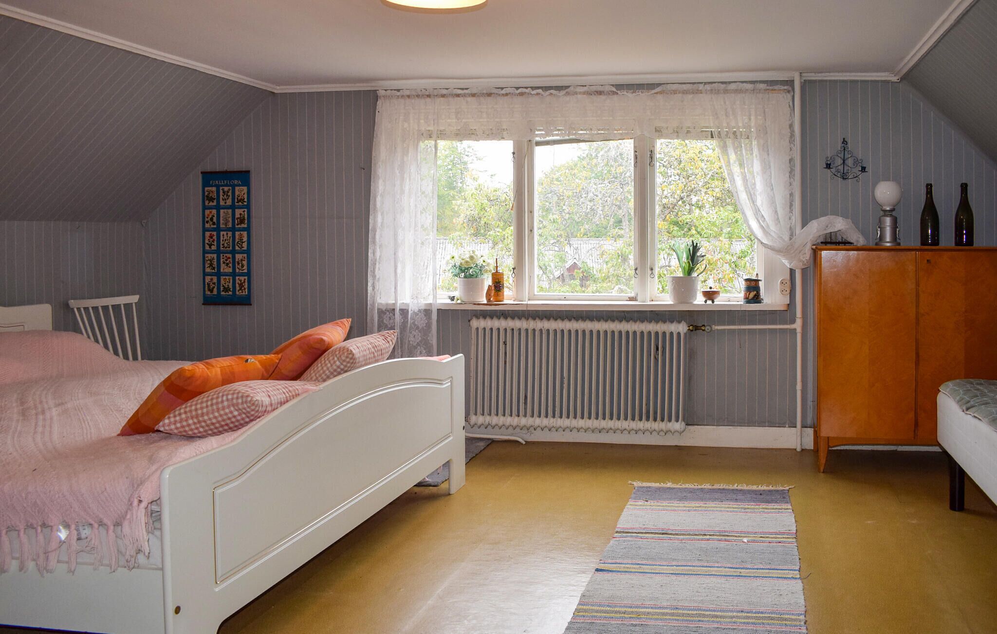 A bedroom with a bed, a radiator, a window with curtains, a wooden cabinet, and a wall-mounted shelf with bottles.