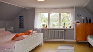Un dormitorio con cama, un radiador, una ventana con cortinas, un armario de madera y una estantería de pared con botellas.