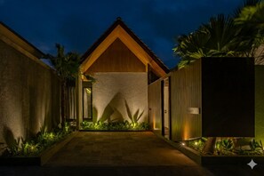 Un edificio moderno con fachada de madera, un camino de hormigón y plantas tropicales.