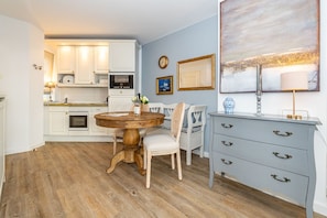 A kitchen with white cabinets, a wooden dining table, and a light blue wall with artwork. - Altstadttor 17 - Thatched-roof vacation apartment for up to two people (Sylt)