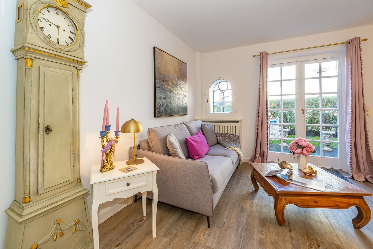 Ein Wohnzimmer mit einer Vintage-Uhr, einer Couch, einem Tisch mit Blumen und einem Fenster mit Vorhängen.