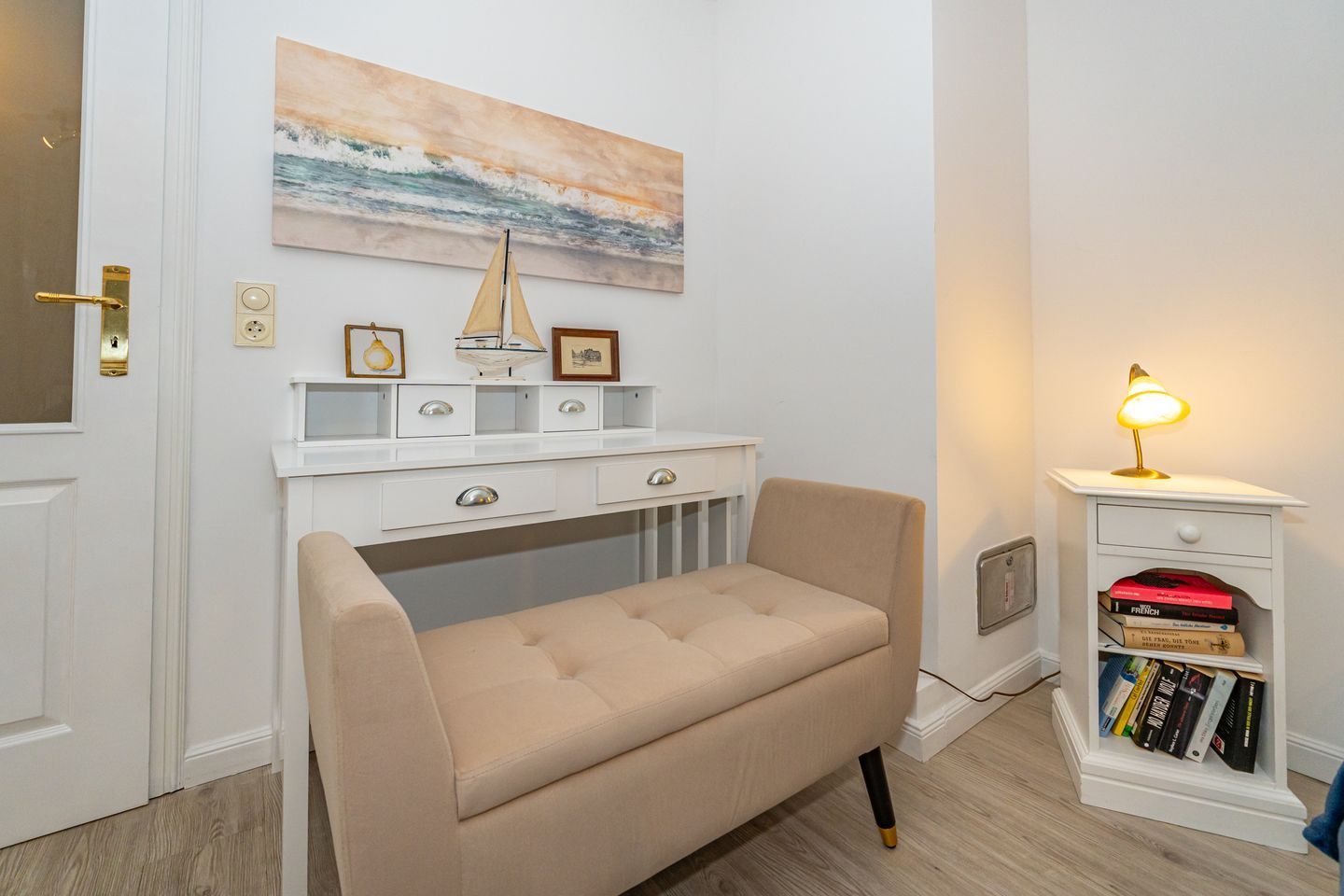 A room with a beige bench, a white desk with drawers, a bookshelf, and a painting of a seascape.