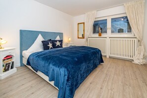 A bedroom with a bed, bedside tables, a window with curtains, a radiator, and a bookshelf. - Altstadttor 17 - Holiday apartment under thatch for up to two people in (Sylt)