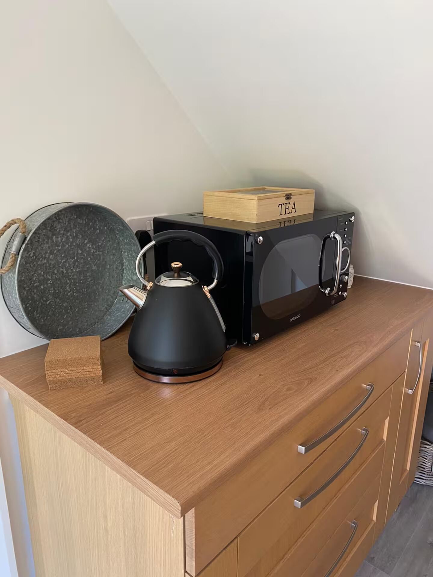 A kitchen with a kettle, a microwave, and a frying pan on a wooden cabinet.