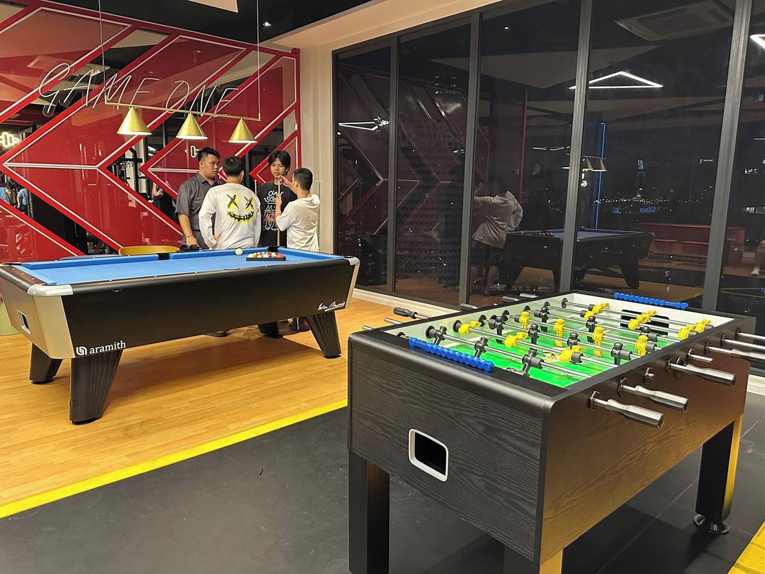 A pool table and a foosball table in a modern indoor setting with large windows and a red geometric wall design.