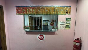A hotel reception area with a pink wall, a fire extinguisher, a window with a decorative curtain, a sign for NIDA rooms, and a price list on the wall. - Hotel No.1 Lodge (Nilai)