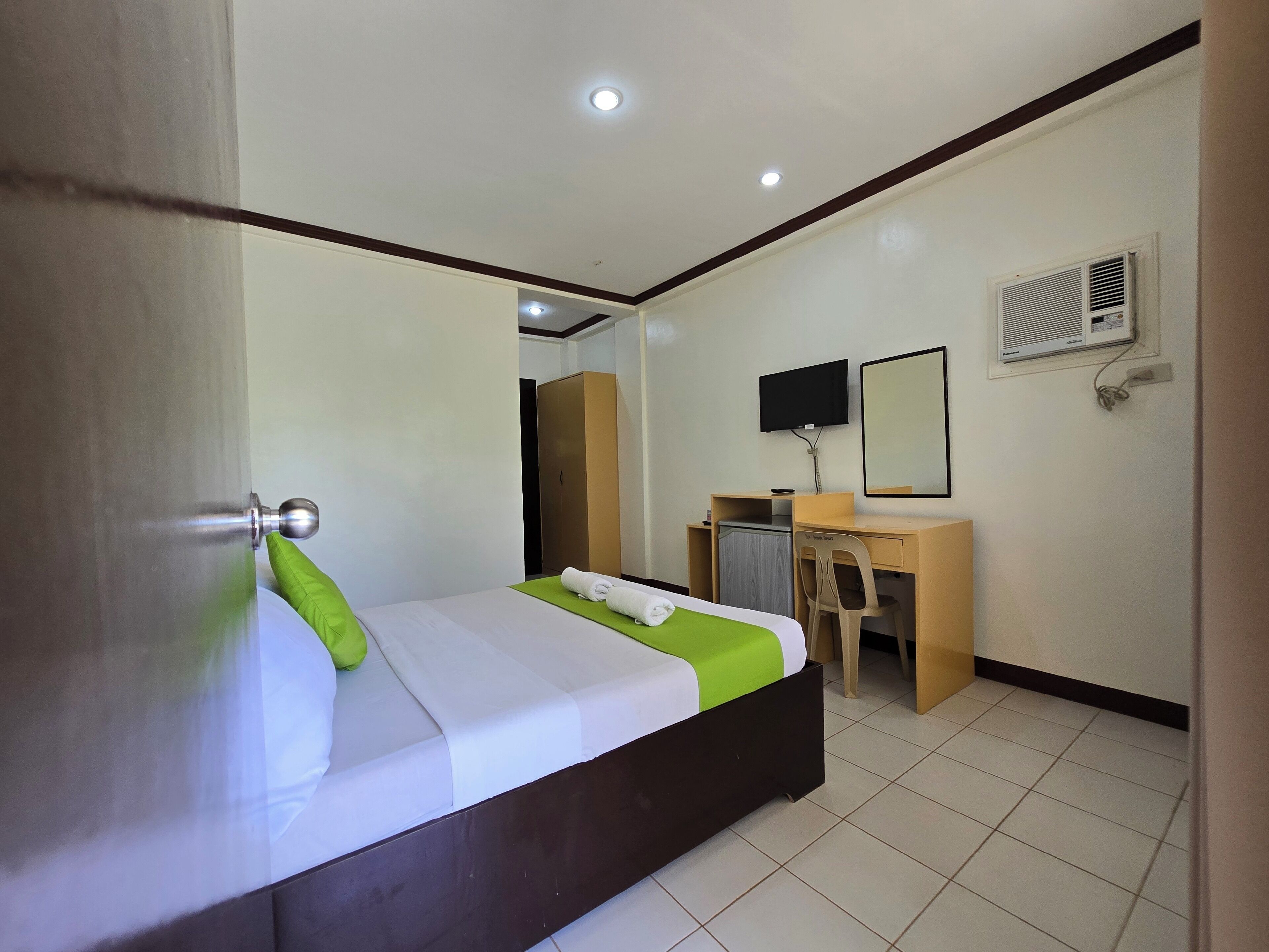 A hotel room with a bed, a desk, a chair, a TV, and an air conditioning unit.